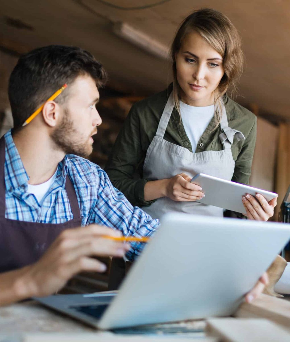 Pretty young carpenter and her bearded colleague brainstorming in workshop: he showing her something on computer screen while she looking at it with interest